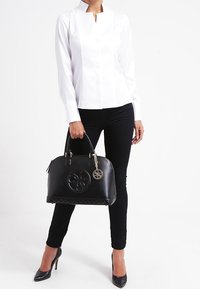 Svart läderhandväska med kviltad struktur, rund logodetalj och guldfärgad hårdvara. Modellen bär en vit skjorta och svarta slim fit-jeans.