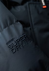 Marineblå pufferjakke med tekstureret stof, udstyret med et broderet "SUPER DRY" logo i mørk tråd på forsiden. Bemærk: minimale designdetaljer.