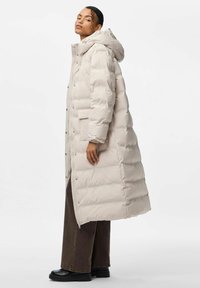 Lunga giacca puffer color beige chiaro con cappuccio, caratterizzata da una texture trapuntata, chiusure a scatto e due tasche laterali. Indossata sopra pantaloni scuri e stivali.