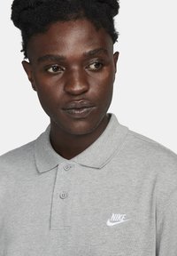 Harmaa polo-paita, joka on valmistettu puuvillasta. Paita sisältää kolme nappia edessä ja pieni valkoinen Nike-logo vasemmassa rinnassa.