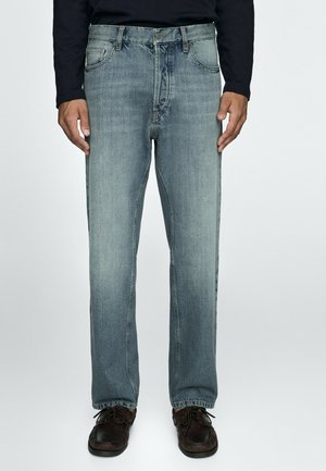 Mand iført falmede blå jeans med lige ben, sort langærmet skjorte og mørkebrune snørestøvler står med hænder afslappet ved siderne.