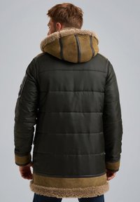 Gevulde donkergroene jas met een met schapenvacht gevoerde capuchon, bruine suède accenten en ribgebreide manchetten. Beschikt over horizontale stiksels en zijzakken.