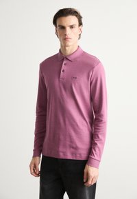 Laza ujjú póló mauve színben, puha anyagból készült, háromgombos kivágással és kis logóval a mellrészen. Sötét farmernadrág viselése.
