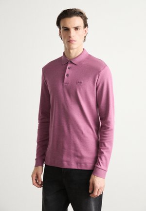 Cămașă polo cu mânecă lungă, de culoare mauve, confecționată dintr-un material moale, având un guler cu trei nasturi și un detaliu mic cu logo pe piept. Se poartă blugi închis la culoare.