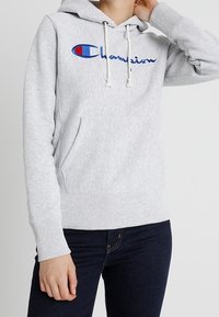Sweat à capuche en molleton gris avec une poche kangourou, cordons ajustables et logo "Champion" brodé en bleu et rouge sur le devant.