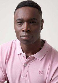 Polo de color rosa claro con cuello, que presenta dos botones y un pequeño logotipo bordado en el lado izquierdo del pecho. Textura de algodón suave.