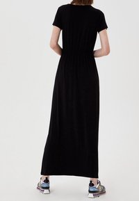 Schwarzes Maxikleid mit kurzen Ärmeln, tailliertem Schnitt und fließendem Design. Kombiniert mit mehrfarbigen Sneakern mit strukturierten Akzenten.