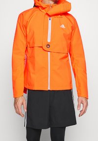 Giacca impermeabile arancione brillante con zip frontale, cappuccio e design pieghevole; presenta dettagli perforati e accenti con il logo.