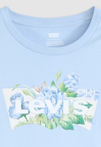 Lyseblå bomull T-skjorte med en blomstergrafikk med blå blomster og grønne blader, sammen med den hvite "Levi's"-logoen.