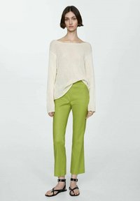 Vrouw in een crèmekleurige gebreide trui, felgroene rechtlopende broek en zwarte sandaaltjes met enkelbandjes, staande tegen een effen achtergrond.