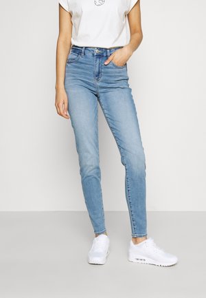 Jeans de tiro alto en azul claro con un corte ajustado, que cuentan con cinco bolsillos y un efecto desgastado. Combinados con una camiseta blanca holgada y zapatillas blancas.