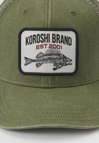 Gorra verde oliva con una parte frontal estructurada, que presenta un parche bordado de un esqueleto de pez y el texto "KOROSHI BRAND EST. 2001."