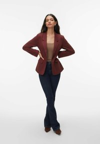 Blazer bordeaux à silhouette ajustée, avec des revers crantés et deux poches avant, assorti à un jean évasé bleu foncé et des chaussures à talons marron.