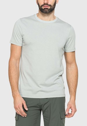 Uomo che indossa una maglietta grigio chiaro a maniche corte con collo a giro e pantaloni cargo verde scuro, in piedi davanti a uno sfondo chiaro uniforme.