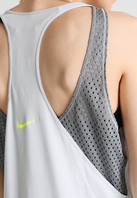 Grijze sportieve tanktop met een ademend mesh-ontwerp, open rug en een klein groen logo op de bovenlinkerzijde.