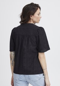 Blusa nera ricamata con maniche a sbuffo corte e schiena arricciata. Caratterizzata da un motivo testurizzato e una vestibilità rilassata, abbinata a jeans azzurri chiari.