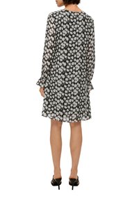 Schwarzes und weißes durchsichtiges Kleid mit gepunktetem Muster, langen Ärmel, weit auslaufenden Manschetten und lockerer Passform, knielang, kombiniert mit schwarzen High Heels.