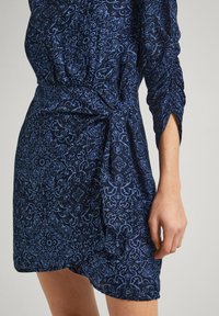 Tmavomodré šaty s prepracovaným svetlomodrým kvetinovým vzorom, krátka sukňa, naťahované rukávy a viazaný pás pre priliehavý siluetu.