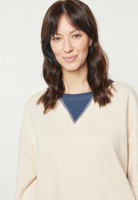 Felpa color crema con colletto blu navy, caratterizzata da un design a forma di V e una vestibilità casual. Texture morbida, con dettagli di cucitura in risalto.