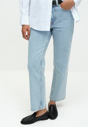 Person wearing light blue straight-leg jeans with a black belt and black slip-on loafers, dressed in a white long-sleeve shirt.