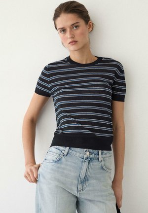 Ung kvinde med håret trukket tilbage iført en sort og blå stribet kortærmet top og lyseblå højtaljede jeans mod hvid baggrund.