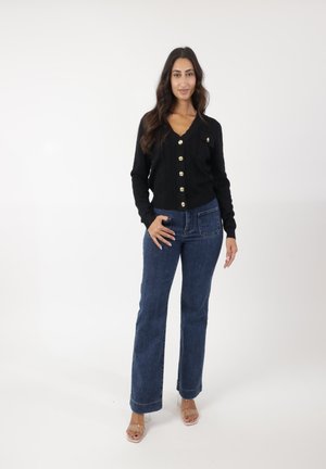 Cardigan noir à côtes avec des boutons dorés, associé à un jean flare bleu taille mi-haute. Des sandales transparentes à talons complètent l'ensemble. Fond blanc.