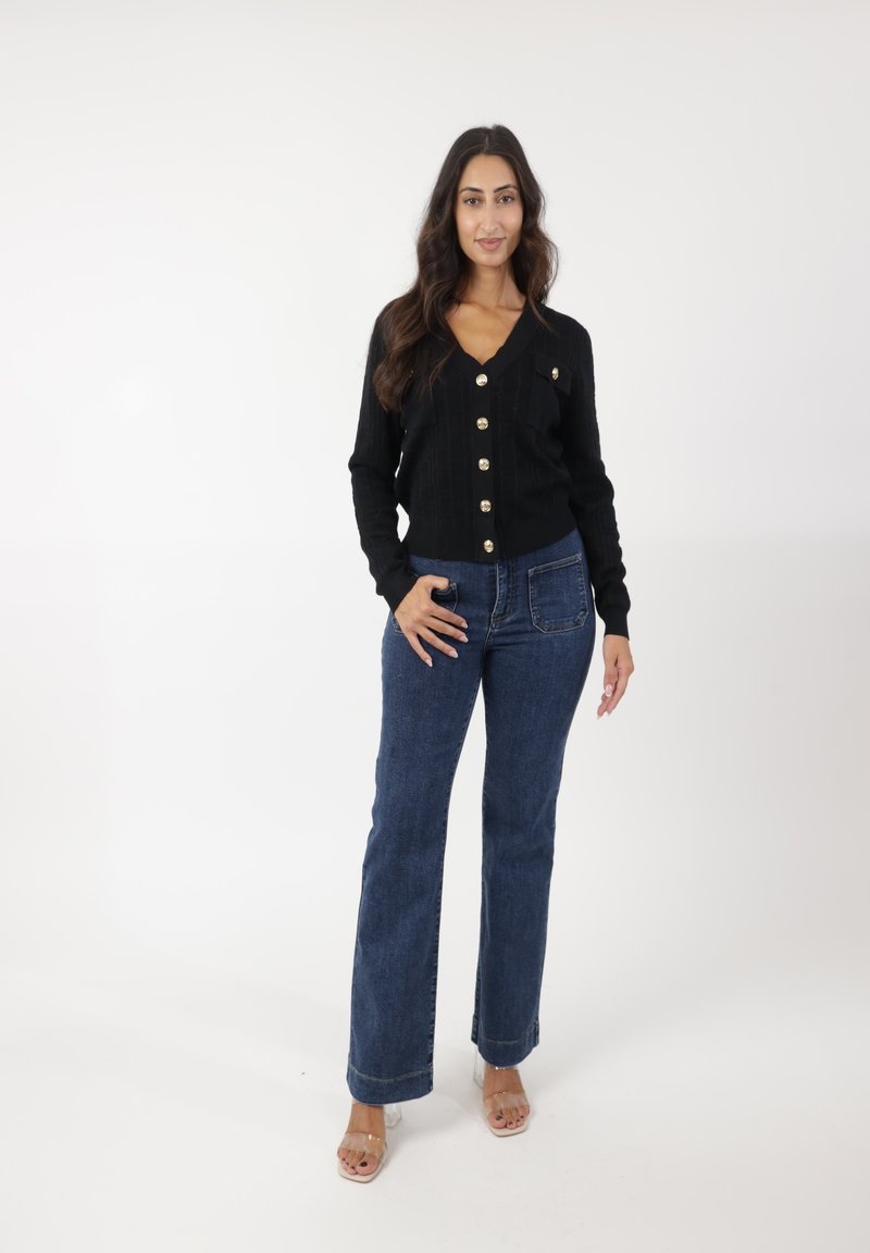 Cárdigan negro de canalé con botones dorados, combinado con jeans acampanados azul de tiro medio. Sandalias de tacón transparente completan el conjunto. Fondo blanco.