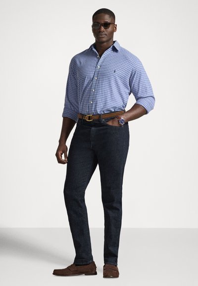 Niebieska koszula w kratę zapinana na guziki z podwiniętymi rękawami, ciemne dżinsy, brązowy pasek oraz brązowe mokasyny, do tego okulary przeciwsłoneczne i zegarek.