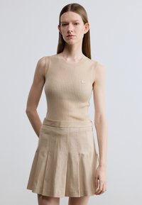 Top sin mangas beige acanalado combinado con una falda plisada beige. La parte superior presenta un cuello redondo y un sutil detalle de logotipo. Textura de tela suave.