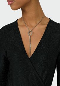 Collar de metal plateado con un lazo retorcido en el centro y un colgante largo y afilado. Llevado con una blusa negra de textura con envoltura.