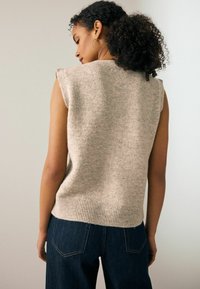 Kortsuurustega naine, kellel on lokkis juuksed, kannab ringikujulisi kõrvarõngaid, beeži värvi ilma varrukateta kudumit ja tumehilse teksaseid, nähtuna seljalt.