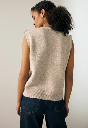 Žena s kučeravými vlasmi, ktorá má na sebe náušnice v tvare kruhu, béžový bezrukávový pletený sveter a tmavomodré džínsy, zobrazená zozadu.