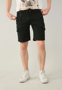 Pantaloni cargo neri realizzati in un tessuto resistente, con tasche laterali, vita con cordino e design a taglio dritto. Indossati con sneaker bianche.