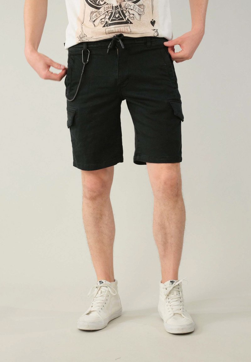 Pantaloni cargo neri realizzati in un tessuto resistente, con tasche laterali, vita con cordino e design a taglio dritto. Indossati con sneaker bianche.