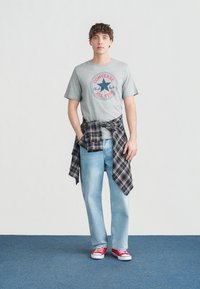 Νέος άνδρας με γκρι μπλουζάκι Converse All Star, ανοιχτόχρωμα τζιν, κόκκινα αθλητικά παπούτσια και μαύρο πουκάμισο με καρό που είναι δεμένο γύρω από τη μέση του, στέκεται σε μπλε πάτωμα.