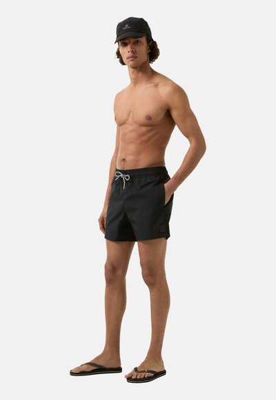 Shorts de bain noirs avec une taille élastique et un cordon de serrage. Le modèle porte des tongs noires et une casquette noire. Tissu lisse, design minimaliste.