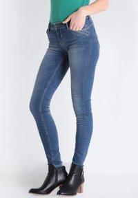 Jeans de denim azul ajustados con un ligero desgaste, cinco bolsillos y cierre con cremallera. Combinados con botines negros de tobillo con un acabado texturizado.