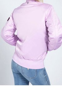 Giacca bomber viola chiaro con una texture lucida, polsini a coste, orlo aderente e un accentuato triangolare sul retro. Abbinata a jeans blu.