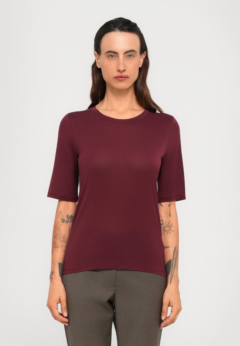 Person with long dark hair, tattoos on both arms, wearing a burgundy short-sleeve top and gray pants, standing against a plain background.