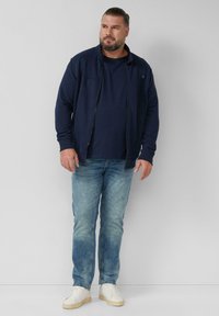 Granatowa kurtka zapinana na zamek z wysokim kołnierzem, połączona z ciemnym t-shirtem, jasnoniebieskimi dżinsami i białymi trampkami. Casualowy strój z minimalnymi detalami.