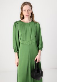 Zelena satenasta haljina dugih rukava s plisiranim detaljima i nabranim manžetama; uz to ide crna torbica s zakrivljenom drškom.