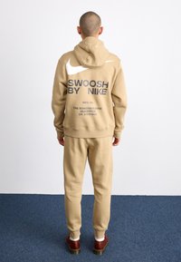 Beige hettegenser og joggebukser sett. Hettegenseren har hvit Nike-logo og teksten "SWOOSH BY NIKE". Stoffet er mykt og glatt, med ribbede mansjetter.