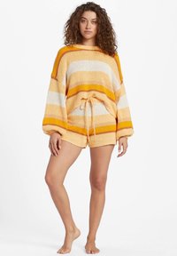 Donna con capelli ricci, che indossa un maglione di maglia a righe largo e pantaloni corti abbinati nei toni di giallo, arancione e crema, in piedi a piedi nudi.