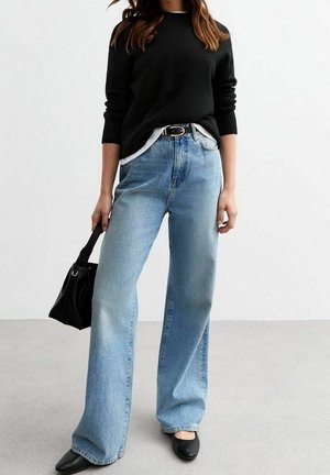 Femme portant un pull noir, un jean large bleu clair, une ceinture noire, des ballerines noires, tenant un sac à main noir sur un fond blanc uni.