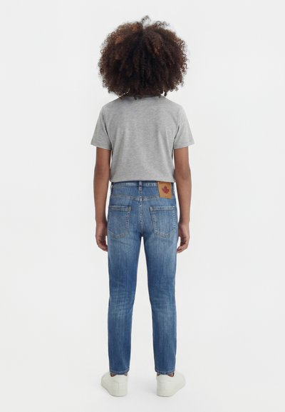 Enfant aux cheveux bouclés portant un t-shirt gris à manches courtes, un jean bleu avec un patch en cuir et des baskets blanches, debout, de dos.