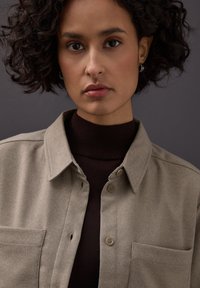 Beiges Hemd mit durchgehendem Knopfverschluss und strukturierter Webart, ausgestattet mit zwei Fronttaschen und einem dunkelbraunen Rollkragenpullover darunter.