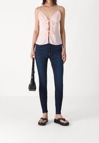 Ljusrosa ärmlös topp med v-ringning och frontknut; tillsammans med mörkblå skinny jeans och svarta flip-flops, håller en liten svart väska.