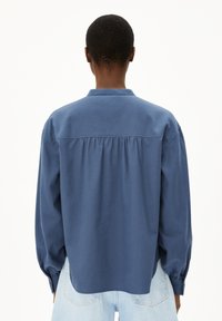 Persona con capelli corti che indossa una camicia blu a maniche lunghe e jeans blu chiaro, vista di schiena su uno sfondo neutro.