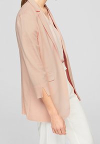 Vaaleanpunainen bleiseri, valmistettu sileästä kankaasta, jossa on räätälöity istuvuus, lovelliset kaulukset ja hillitty halkio hihoissa.