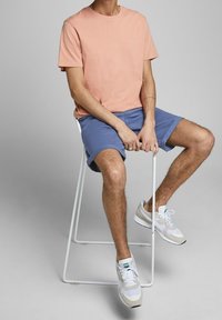 Světle růžové bavlněné tričko, modré šortky a bílé tenisky se šedými detaily, sedící na bílé stoličce proti šedému pozadí.